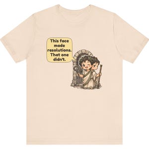 K&ouml;nnte beinhalten: Ein hellbeiges T-Shirt mit einer Grafik einer zweigesichtigen Figur, die einen Lorbeerkranz und eine Toga tr&auml;gt. Ein Gesicht zwinkert und h&auml;lt einen Schl&uuml;ssel, das andere schaut ernst und h&auml;lt einen Stab. Eine Sprechblase enth&auml;lt den Text: This face made resolutions. That one didn't.