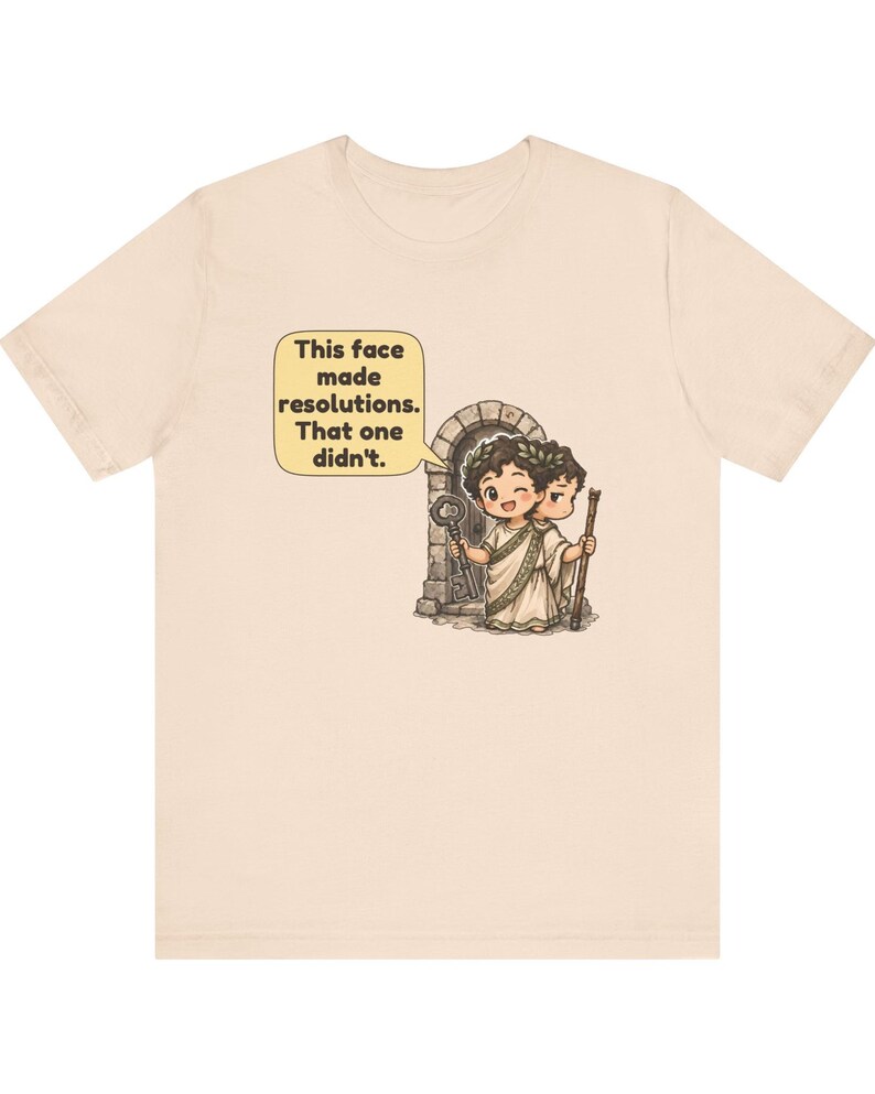 K&ouml;nnte beinhalten: Ein hellbeiges T-Shirt mit einer Grafik einer zweigesichtigen Figur, die einen Lorbeerkranz und eine Toga tr&auml;gt. Ein Gesicht zwinkert und h&auml;lt einen Schl&uuml;ssel, das andere schaut ernst und h&auml;lt einen Stab. Eine Sprechblase enth&auml;lt den Text: This face made resolutions. That one didn't.