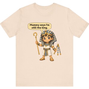 Może przedstawiać: Jasnobeżowy t-shirt z rysunkową ilustracją młodego faraona w niebiesko-złotym nakryciu głowy, białej tunice i sandałach. Postać trzyma złote berło i bicz, a nad nią znajduje się dymek z napisem: Mummy says I'm still the king.