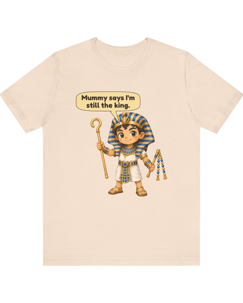 Może przedstawiać: Jasnobeżowy t-shirt z rysunkową ilustracją młodego faraona w niebiesko-złotym nakryciu głowy, białej tunice i sandałach. Postać trzyma złote berło i bicz, a nad nią znajduje się dymek z napisem: Mummy says I'm still the king.