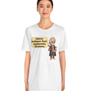 Pu&ograve; includere: Una t-shirt bianca con un'illustrazione a cartoni animati di un uomo con gilet marrone e cappotto scuro che tiene in mano una penna e una pergamena. Un fumetto giallo contiene il testo: Hebrew professor. Poet. Accidentally Christmas.