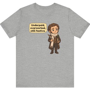 May include: A heather gray t-shirt featuring a cartoon illustration of a person in a brown coat and white scarf holding a quill and candle. A speech bubble next to the character contains the text: Underpaid, overworked, still festive.