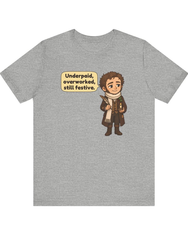 May include: A heather gray t-shirt featuring a cartoon illustration of a person in a brown coat and white scarf holding a quill and candle. A speech bubble next to the character contains the text: Underpaid, overworked, still festive.