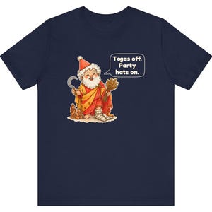 May include: A navy blue t-shirt featuring a graphic of a bearded figure wearing a red party hat and a yellow robe. The figure holds a sickle and a bundle of wheat, with a speech bubble that reads: Togas off. Party hats on.