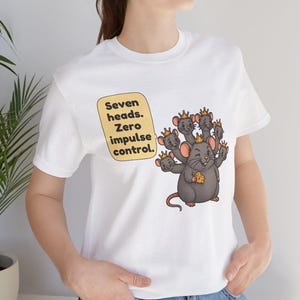 May include: A white t-shirt featuring a graphic of a gray rat with seven heads, each wearing a small gold crown. The rat holds a piece of yellow cheese. A yellow speech bubble contains the black text: Seven heads. Zero impulse control.
