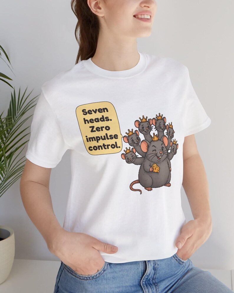 May include: A white t-shirt featuring a graphic of a gray rat with seven heads, each wearing a small gold crown. The rat holds a piece of yellow cheese. A yellow speech bubble contains the black text: Seven heads. Zero impulse control.