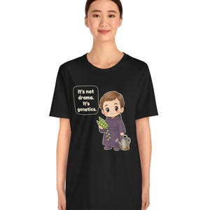 May include: A black t-shirt featuring a cartoon illustration of a child in a purple robe holding a pea pod and a watering can. A speech bubble next to the child contains the text: It's not drama. It's genetics. The design is centered on the chest.