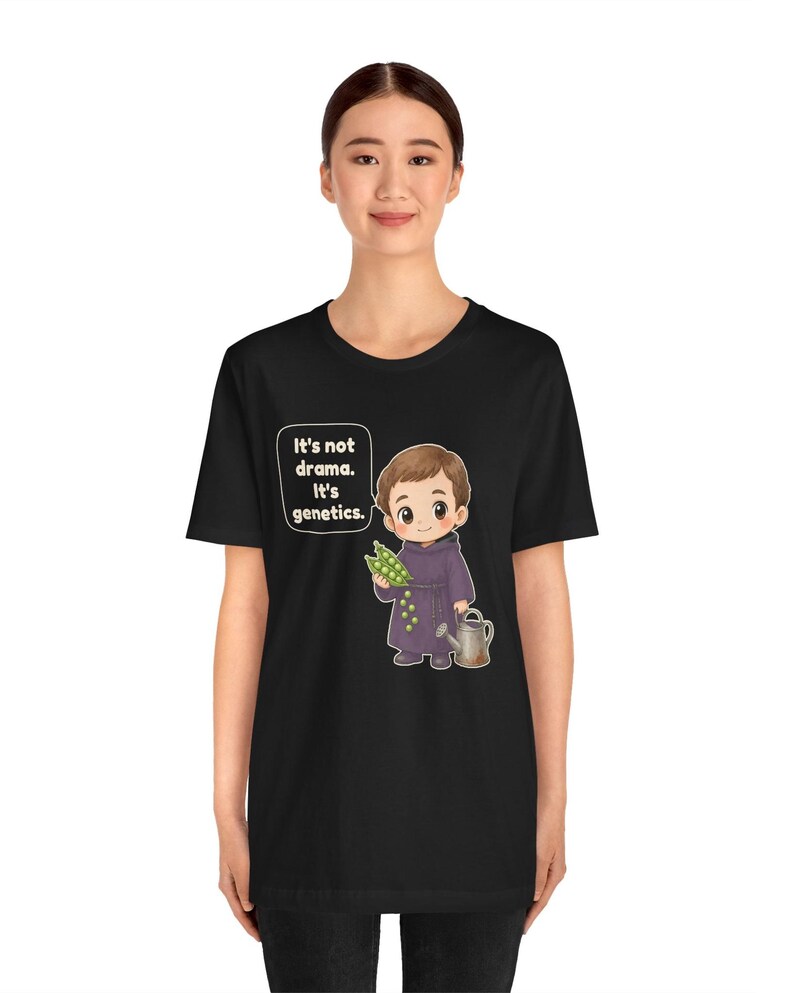 May include: A black t-shirt featuring a cartoon illustration of a child in a purple robe holding a pea pod and a watering can. A speech bubble next to the child contains the text: It's not drama. It's genetics. The design is centered on the chest.