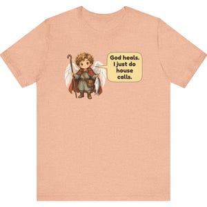 May include: A peach-coloured t-shirt featuring a graphic of a small angel with wings, holding a wooden staff and a fish. A speech bubble next to the angel contains the text: God heals. I just do house calls. The design is centred on the chest.