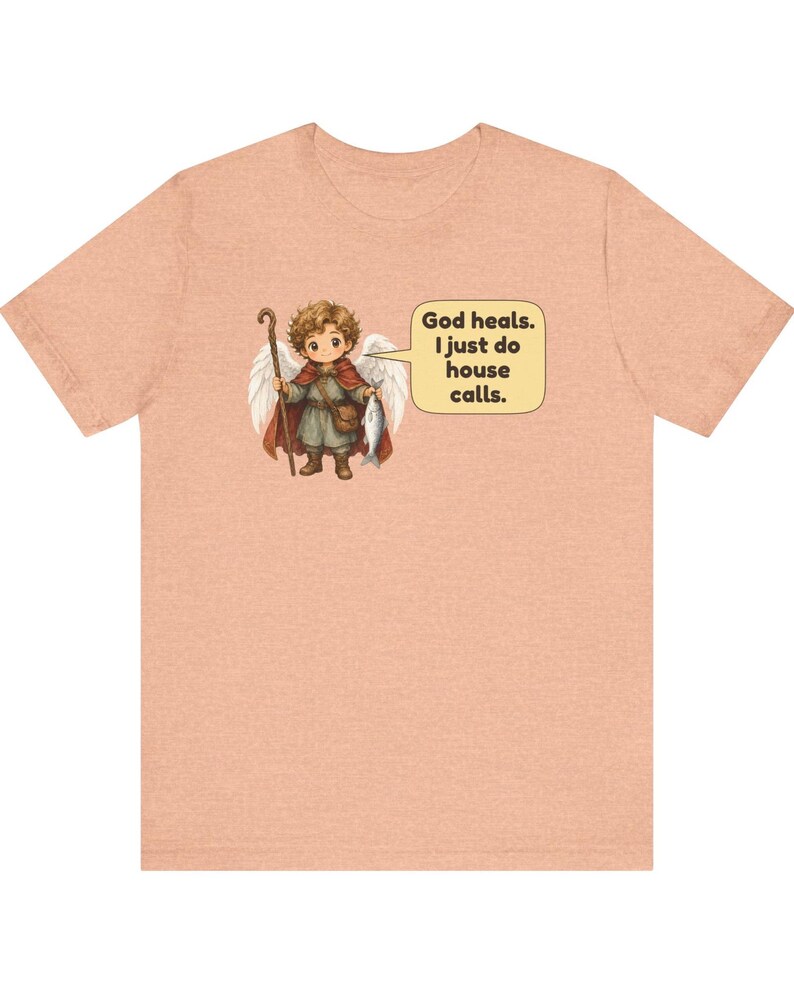 May include: A peach-coloured t-shirt featuring a graphic of a small angel with wings, holding a wooden staff and a fish. A speech bubble next to the angel contains the text: God heals. I just do house calls. The design is centred on the chest.
