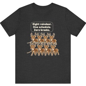 K&ouml;nnte beinhalten: Ein dunkelgraues T-Shirt mit einer Grafik von acht Cartoon-Rentieren, die in zwei Reihen angeordnet sind. &Uuml;ber den Rentieren befindet sich eine wei&szlig;e Sprechblase mit dem Text: Eight reindeer. One schedule. Zero breaks. Die Rentiere sind braun mit beigen Geweihen und Geschirren.