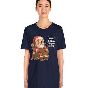 Op de afbeelding: Een marineblauw t-shirt met korte mouwen en een afbeelding van de Kerstman die in een groene fauteuil zit en een boek leest. Een tekstballon ernaast bevat de tekst: Ho ho hold on. Chapter ending. Het ontwerp bevat ook een klein bijzettafeltje.