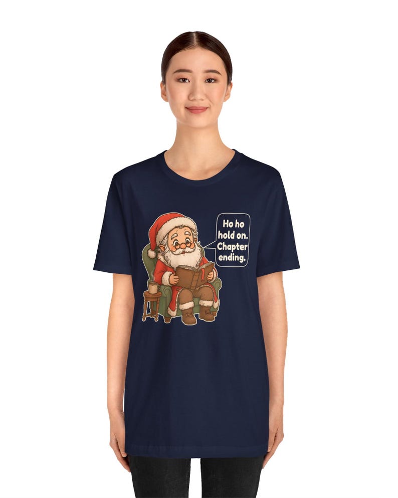 Op de afbeelding: Een marineblauw t-shirt met korte mouwen en een afbeelding van de Kerstman die in een groene fauteuil zit en een boek leest. Een tekstballon ernaast bevat de tekst: Ho ho hold on. Chapter ending. Het ontwerp bevat ook een klein bijzettafeltje.