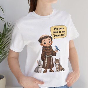 May include: A white t-shirt featuring a cartoon illustration of a monk in a brown robe with a rabbit, a wolf, and a blue bird. A yellow speech bubble above the monk contains the text: My pets talk to me. Yours too?