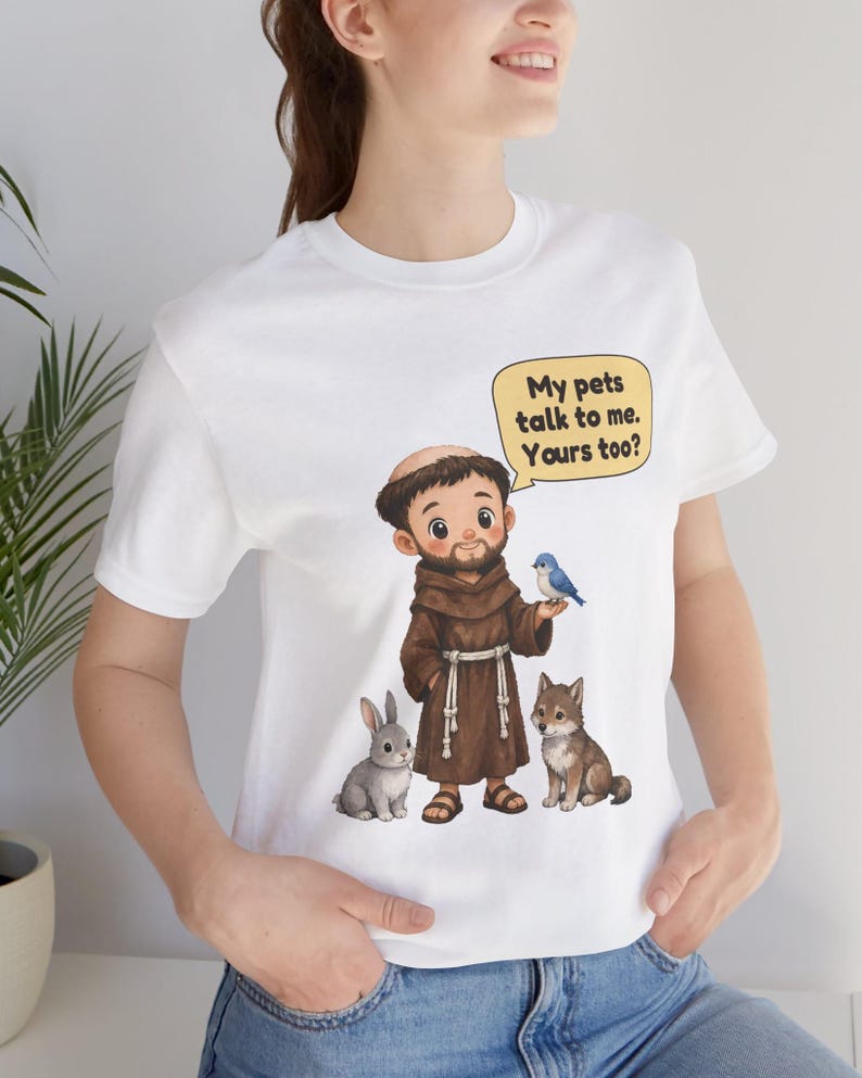 May include: A white t-shirt featuring a cartoon illustration of a monk in a brown robe with a rabbit, a wolf, and a blue bird. A yellow speech bubble above the monk contains the text: My pets talk to me. Yours too?