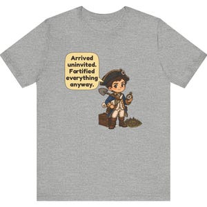 May include: A heather gray t-shirt featuring a cartoon illustration of a colonial-era explorer holding a shovel and compass, standing next to a wooden chest. A speech bubble contains the text: Arrived uninvited. Fortified everything anyway.