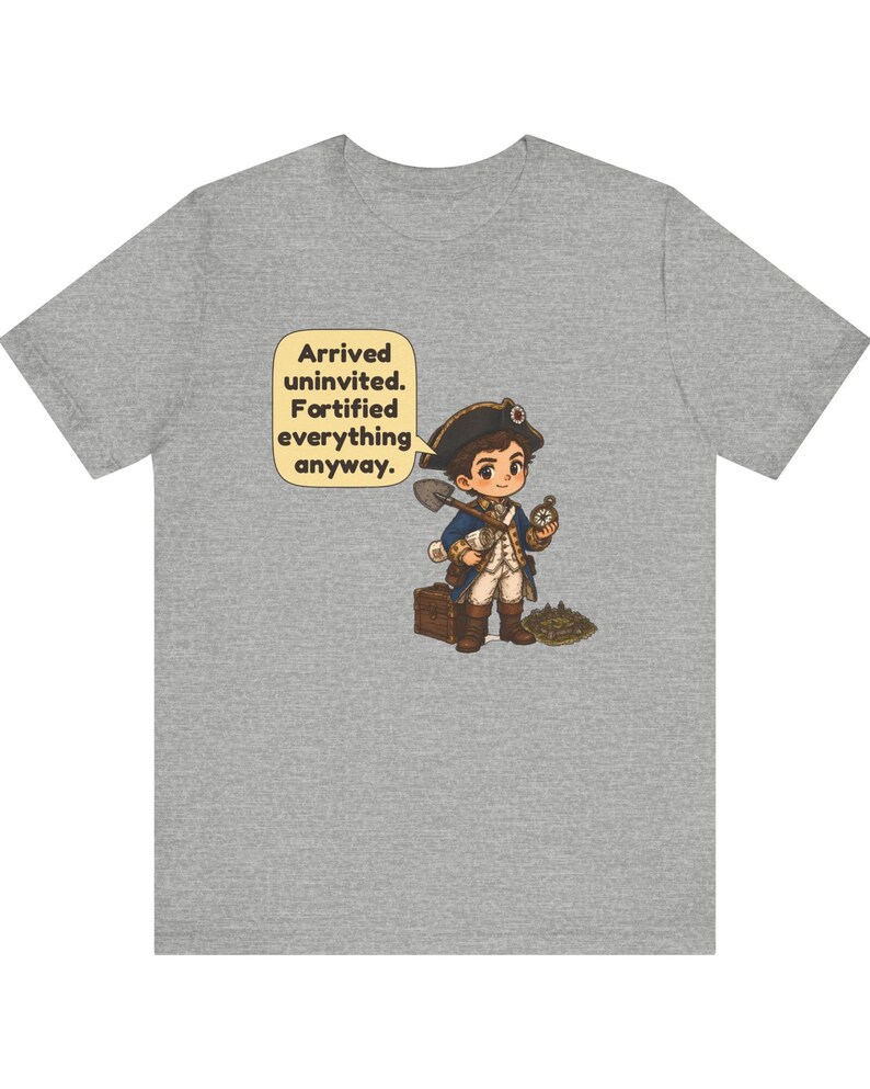 May include: A heather gray t-shirt featuring a cartoon illustration of a colonial-era explorer holding a shovel and compass, standing next to a wooden chest. A speech bubble contains the text: Arrived uninvited. Fortified everything anyway.