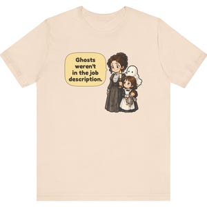 Op de afbeelding: Een lichtbeige t-shirt met een afbeelding van een persoon in een donkere jurk met een kaars, een kind met een pop en een klein wit spookje. Een tekstballon bevat de tekst: Ghosts weren't in the job description.