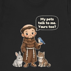 May include: A black t-shirt featuring a cartoon illustration of a bearded figure in a brown robe with a rope belt, standing with a grey rabbit, a small wolf, and a blue bird. A speech bubble contains the text: My pets talk to me. Yours too?