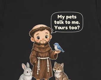 May include: A black t-shirt featuring a cartoon illustration of a bearded figure in a brown robe with a rope belt, standing with a grey rabbit, a small wolf, and a blue bird. A speech bubble contains the text: My pets talk to me. Yours too?