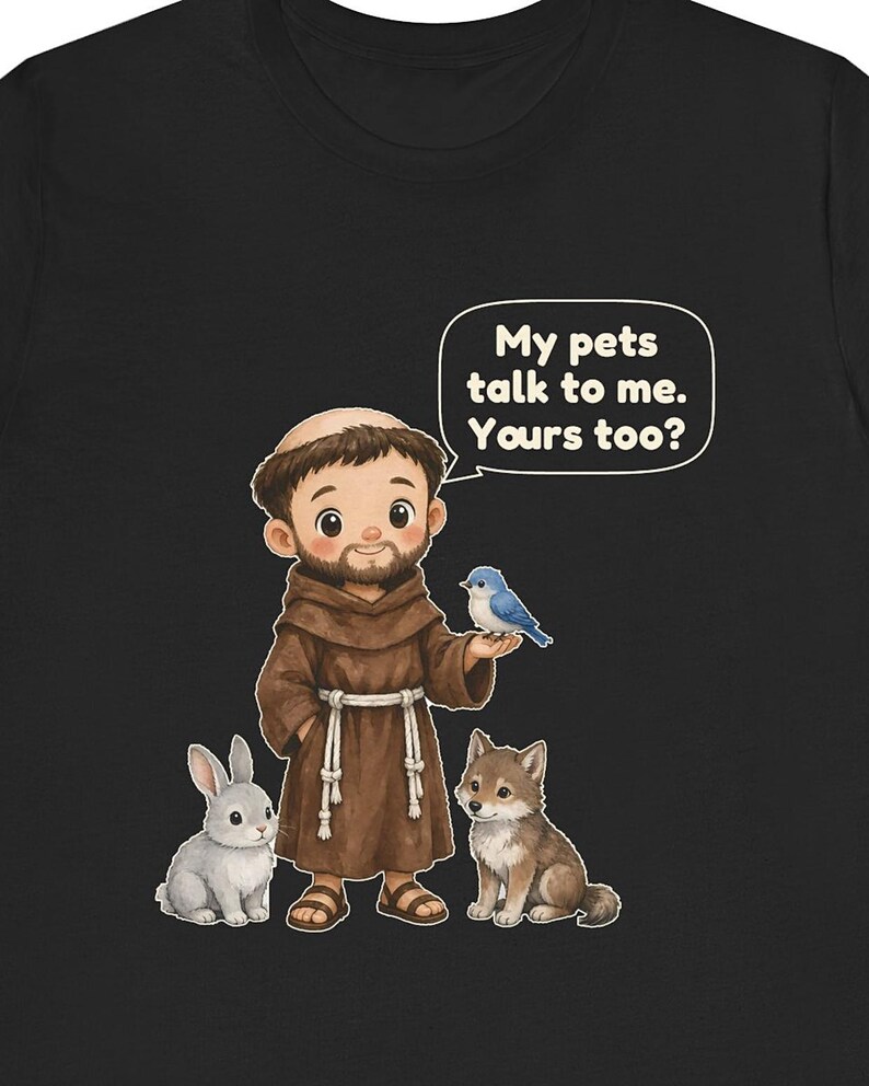 May include: A black t-shirt featuring a cartoon illustration of a bearded figure in a brown robe with a rope belt, standing with a grey rabbit, a small wolf, and a blue bird. A speech bubble contains the text: My pets talk to me. Yours too?