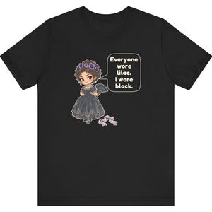 May include: A black t-shirt featuring a cartoon illustration of a character in a black dress with a lilac floral headpiece and a fan. A speech bubble contains the text: Everyone wore lilac. I wore black. A small lilac ribbon lies below.