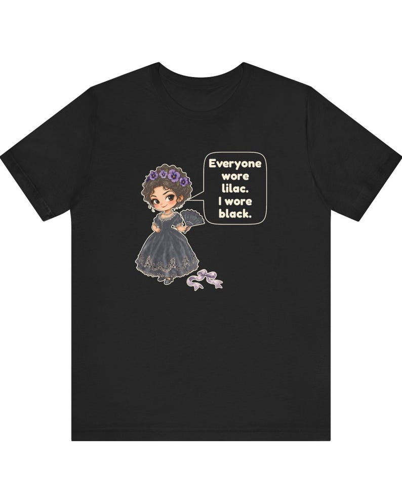 May include: A black t-shirt featuring a cartoon illustration of a character in a black dress with a lilac floral headpiece and a fan. A speech bubble contains the text: Everyone wore lilac. I wore black. A small lilac ribbon lies below.