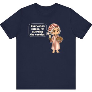 Op de afbeelding: Een marineblauw t-shirt met een stripfiguur in een rood-wit gestreepte pyjama en een bijpassende slaapmuts. Het figuur houdt een brandende kaars en een bord met koekjes vast, met een tekstballon: Everyone's asleep. I'm guarding the cookies.