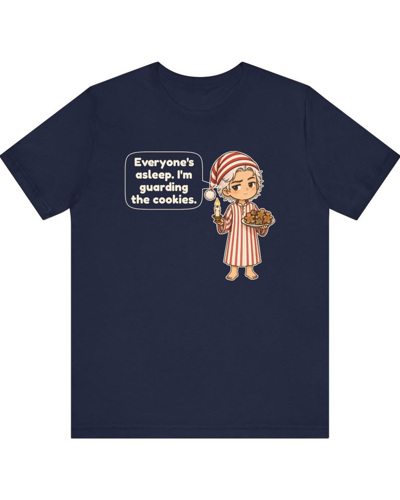 Op de afbeelding: Een marineblauw t-shirt met een stripfiguur in een rood-wit gestreepte pyjama en een bijpassende slaapmuts. Het figuur houdt een brandende kaars en een bord met koekjes vast, met een tekstballon: Everyone's asleep. I'm guarding the cookies.