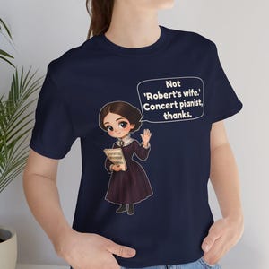 Puede incluir: Una camiseta azul marino con una ilustraci&oacute;n de estilo dibujo animado de una persona con un vestido morado oscuro sosteniendo partituras. Un globo de texto sobre la figura contiene: 'Not 'Robert's wife.' Concert pianist, thanks.'
