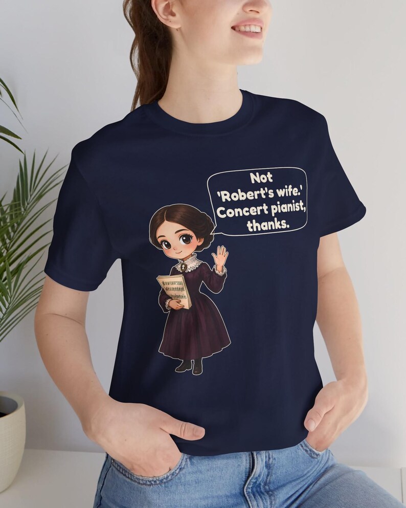 Puede incluir: Una camiseta azul marino con una ilustraci&oacute;n de estilo dibujo animado de una persona con un vestido morado oscuro sosteniendo partituras. Un globo de texto sobre la figura contiene: 'Not 'Robert's wife.' Concert pianist, thanks.'