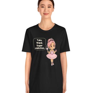 May include: A black t-shirt featuring a graphic of a cartoon ballerina with pink hair, a pink tutu, and a crown, holding a star wand. A speech bubble next to the character contains the text: Tutu. Wand. Sugar addiction.