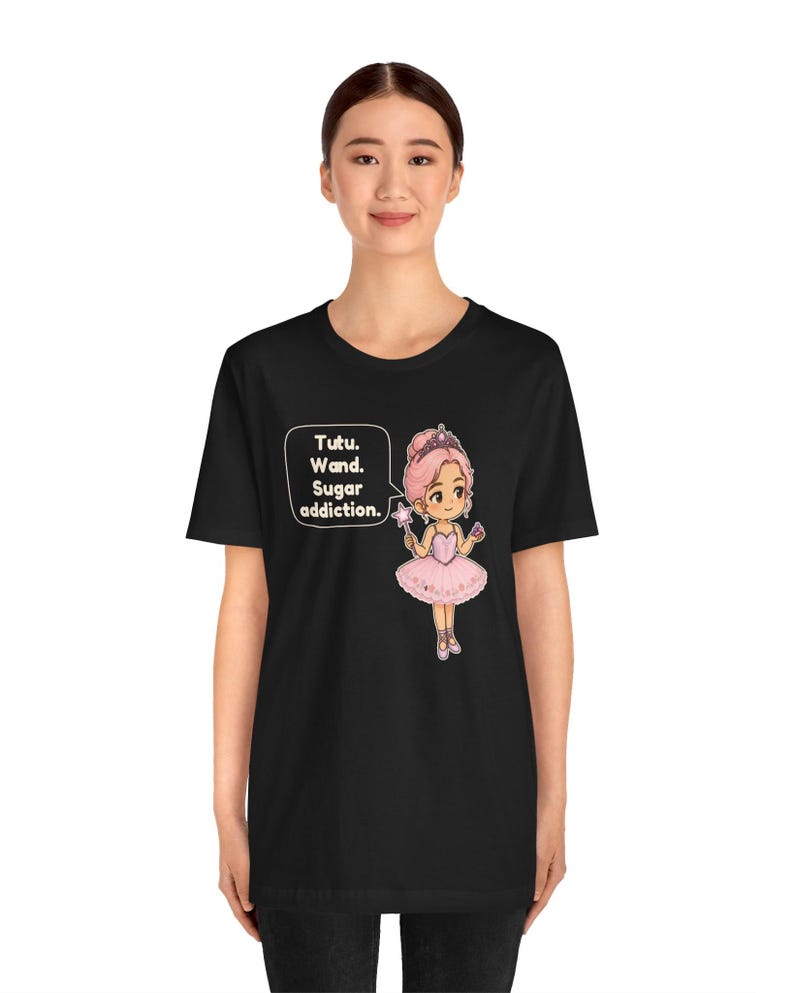May include: A black t-shirt featuring a graphic of a cartoon ballerina with pink hair, a pink tutu, and a crown, holding a star wand. A speech bubble next to the character contains the text: Tutu. Wand. Sugar addiction.