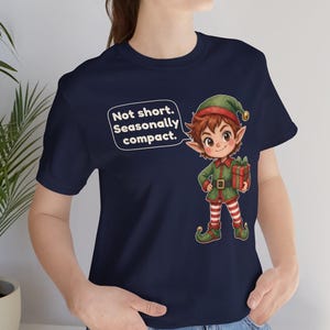 May include: A navy blue t-shirt featuring a graphic of a cartoon elf wearing a green outfit and red and white striped leggings, holding a gift. A speech bubble next to the elf contains the text: Not short. Seasonally compact.