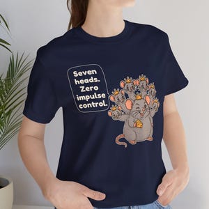 May include: A navy blue short-sleeve t-shirt featuring a graphic of a gray mouse with seven heads, each wearing a small gold crown. The mouse holds a piece of yellow cheese. The text reads: Seven heads. Zero impulse control.