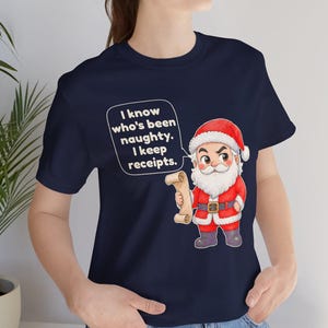 May include: A navy blue t-shirt featuring a cartoon Santa Claus holding a scroll. The text in a speech bubble reads: I know who's been naughty. I keep receipts. The Santa character wears a red suit with white trim, a black belt, and dark boots.