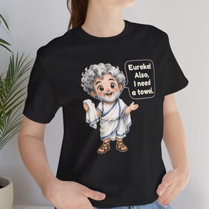 May include: A black t-shirt featuring a cartoon illustration of a bearded man with curly gray hair wearing a white toga with blue trim and brown sandals. The character holds a white towel and a speech bubble that reads, Eureka! Also, I need a towel.