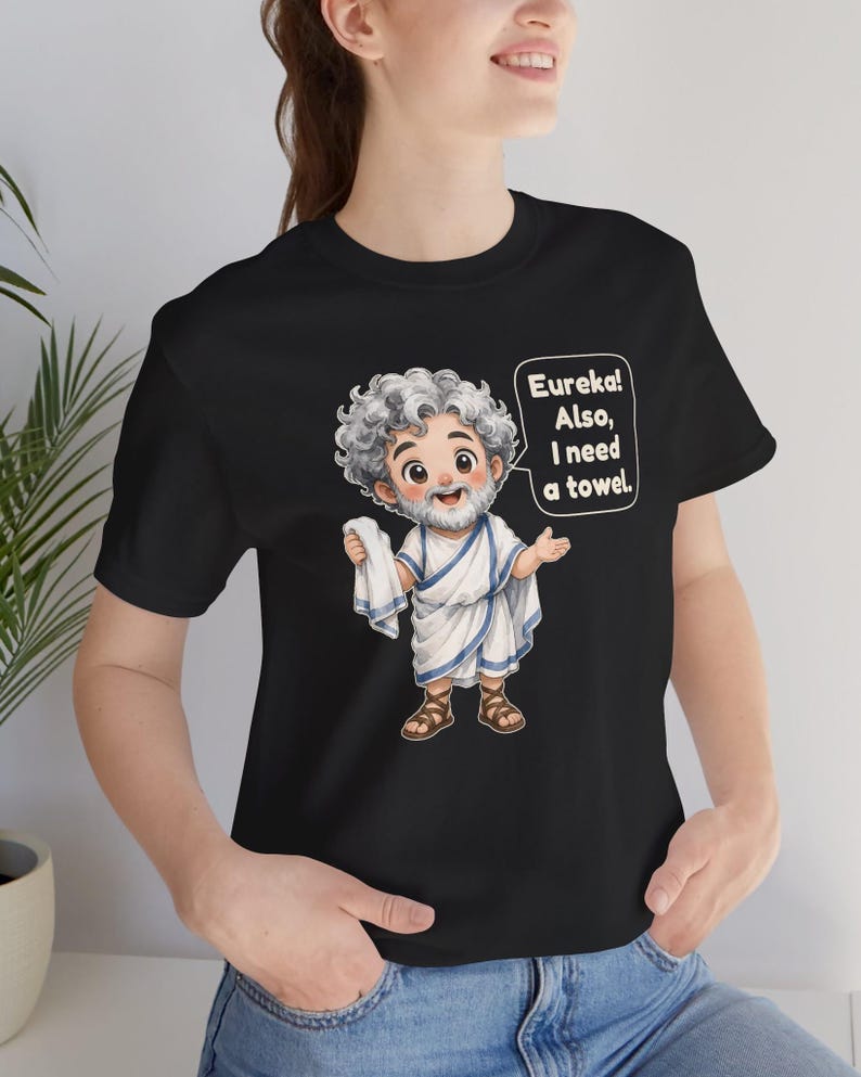 May include: A black t-shirt featuring a cartoon illustration of a bearded man with curly gray hair wearing a white toga with blue trim and brown sandals. The character holds a white towel and a speech bubble that reads, Eureka! Also, I need a towel.
