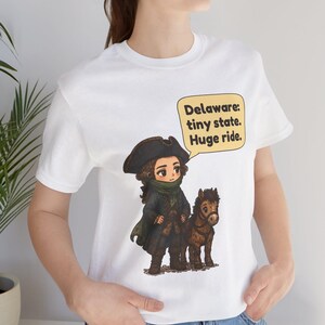 May include: A white t-shirt featuring a cartoon illustration of a historical figure in a tricorn hat and dark coat standing next to a small brown horse. A yellow speech bubble contains the text: Delaware: tiny state. Huge ride.