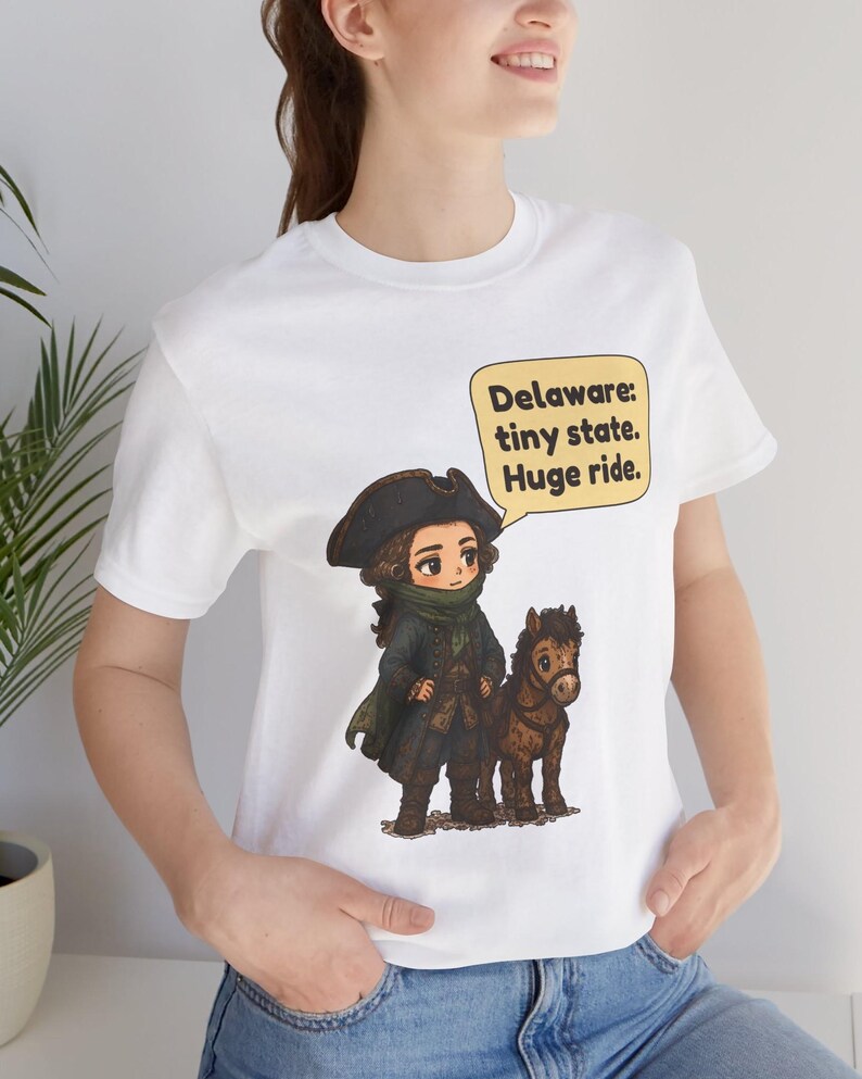May include: A white t-shirt featuring a cartoon illustration of a historical figure in a tricorn hat and dark coat standing next to a small brown horse. A yellow speech bubble contains the text: Delaware: tiny state. Huge ride.