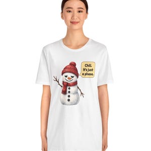 Pode incluir: Uma t-shirt branca de manga curta com o gr&aacute;fico de um boneco de neve a usar um gorro e um cachecol de malha vermelhos. O boneco de neve tem um nariz de cenoura, bra&ccedil;os de ramo e um bal&atilde;o com o texto: Chill. It's just a phase.
