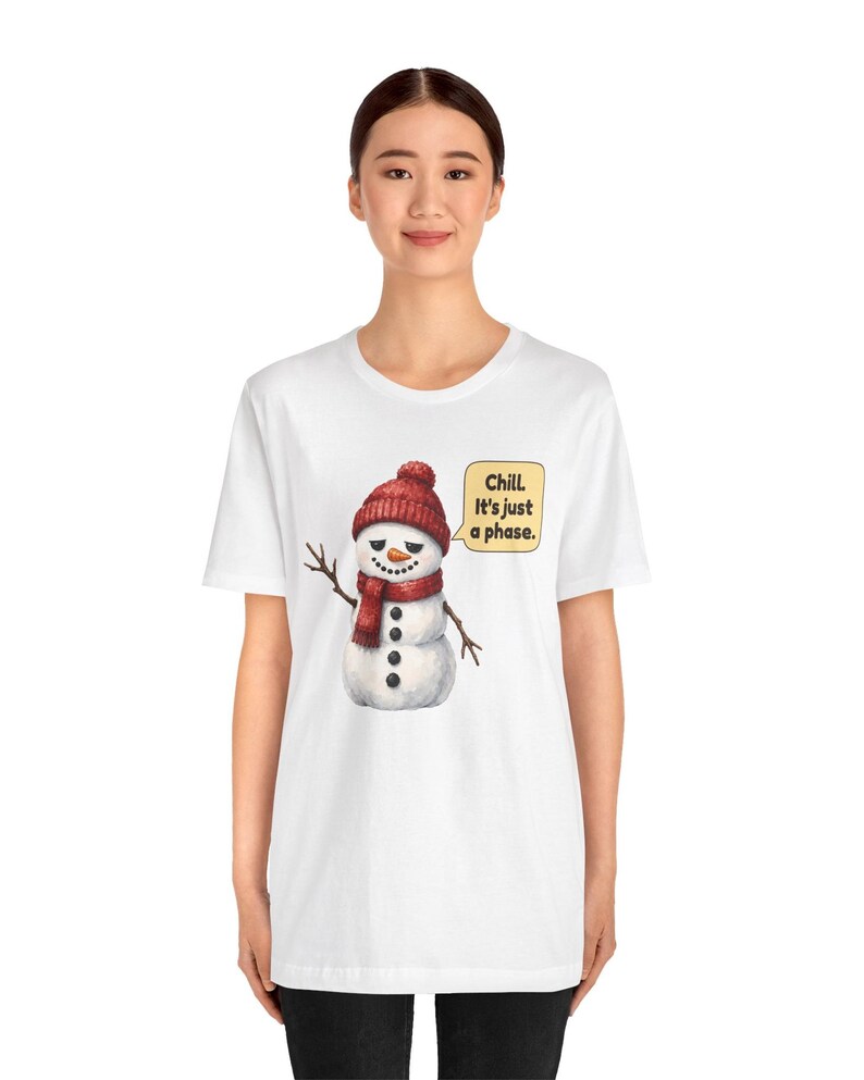 Pode incluir: Uma t-shirt branca de manga curta com o gr&aacute;fico de um boneco de neve a usar um gorro e um cachecol de malha vermelhos. O boneco de neve tem um nariz de cenoura, bra&ccedil;os de ramo e um bal&atilde;o com o texto: Chill. It's just a phase.