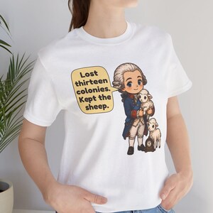 Peut inclure: Un t-shirt blanc avec une illustration de style dessin anim&eacute; repr&eacute;sentant un personnage historique en manteau bleu et perruque blanche tenant un petit mouton blanc, avec un autre mouton &agrave; c&ocirc;t&eacute;. Une bulle contient le texte : Lost thirteen colonies. Kept the sheep.
