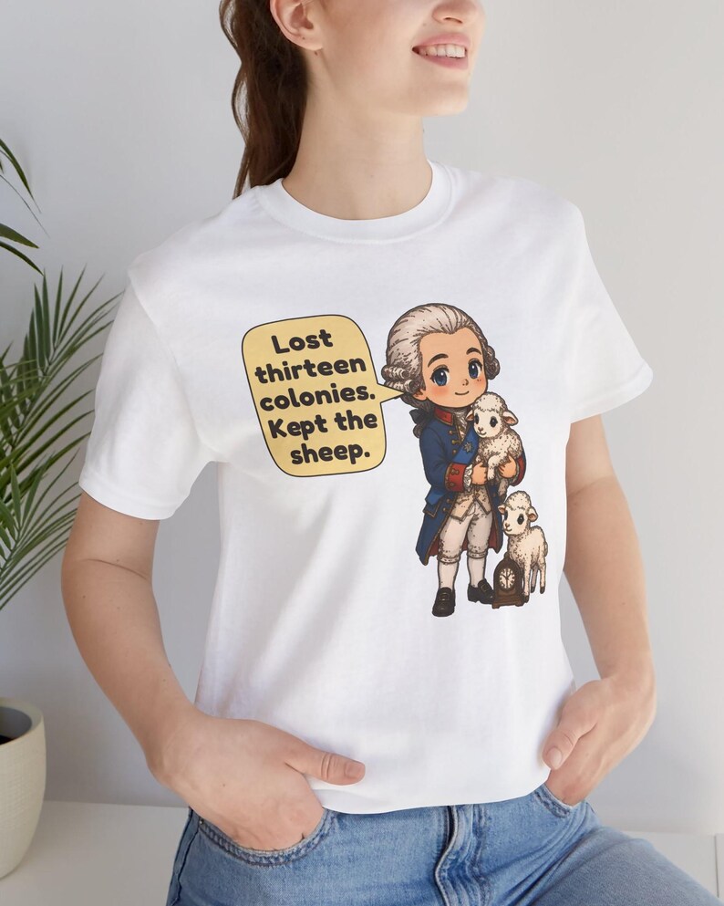Peut inclure: Un t-shirt blanc avec une illustration de style dessin anim&eacute; repr&eacute;sentant un personnage historique en manteau bleu et perruque blanche tenant un petit mouton blanc, avec un autre mouton &agrave; c&ocirc;t&eacute;. Une bulle contient le texte : Lost thirteen colonies. Kept the sheep.