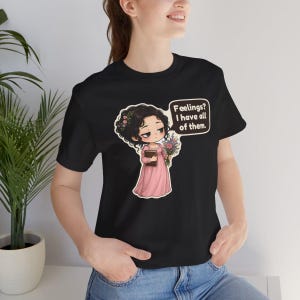 K&ouml;nnte beinhalten: Ein schwarzes Kurzarm-T-Shirt mit einer Cartoon-Illustration einer Person mit dunklem lockigem Haar in einem rosa Kleid, die ein Buch und Blumen h&auml;lt, mit einer Sprechblase: "Feelings? I have all of them."