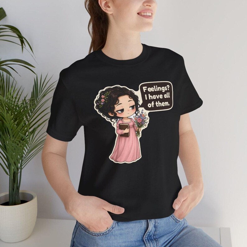 K&ouml;nnte beinhalten: Ein schwarzes Kurzarm-T-Shirt mit einer Cartoon-Illustration einer Person mit dunklem lockigem Haar in einem rosa Kleid, die ein Buch und Blumen h&auml;lt, mit einer Sprechblase: "Feelings? I have all of them."