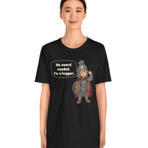 May include: A black t-shirt featuring a cartoon illustration of a warrior in chainmail and a helmet, holding a round shield. A speech bubble next to the character contains the text: No sword needed. I'm a hugger.