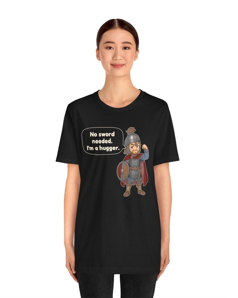May include: A black t-shirt featuring a cartoon illustration of a warrior in chainmail and a helmet, holding a round shield. A speech bubble next to the character contains the text: No sword needed. I'm a hugger.