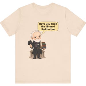 May include: A light beige t-shirt featuring a cartoon illustration of an elderly man with a white beard, wearing a black coat and scarf, holding a stack of books. A speech bubble contains the text: Have you tried the library? I built a few.