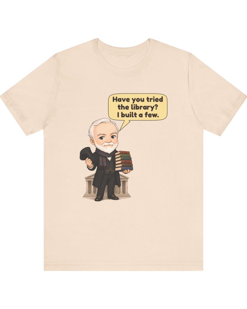 May include: A light beige t-shirt featuring a cartoon illustration of an elderly man with a white beard, wearing a black coat and scarf, holding a stack of books. A speech bubble contains the text: Have you tried the library? I built a few.